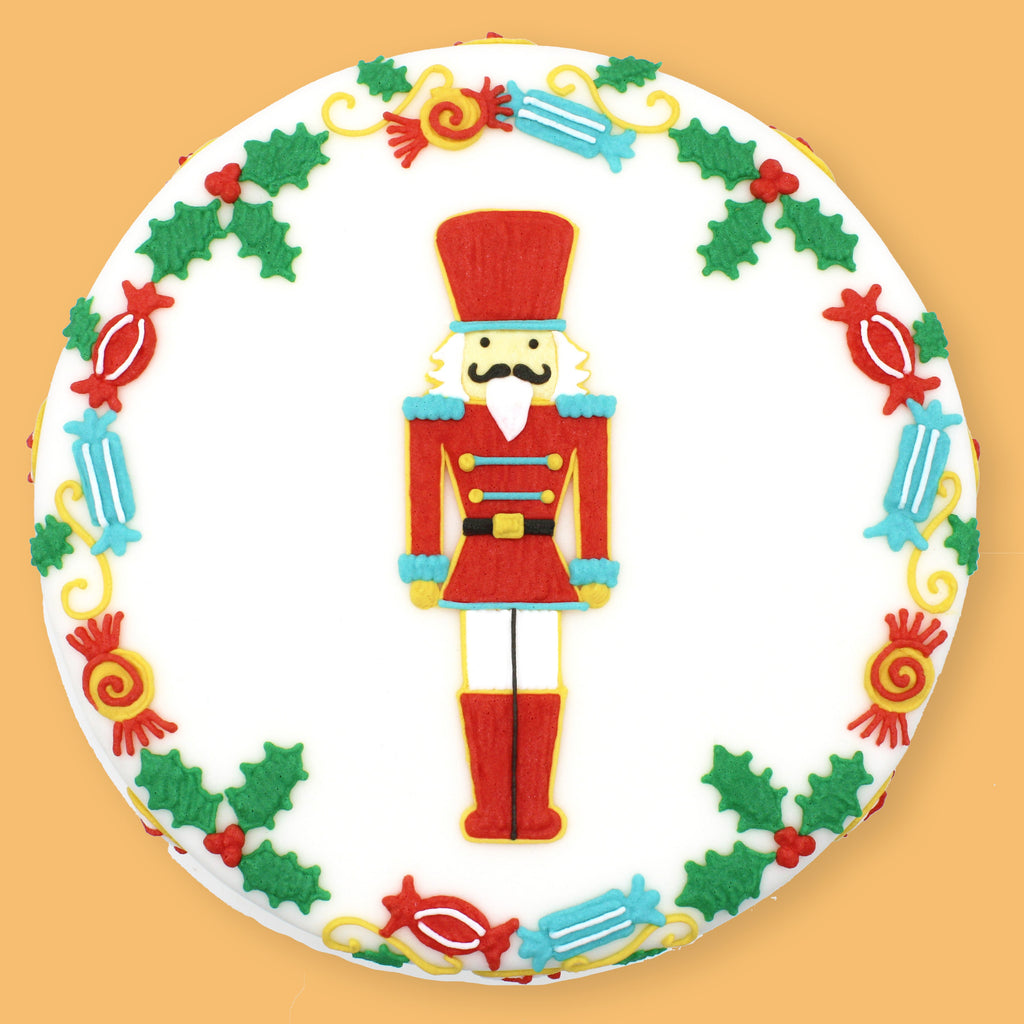 A round white cake decorated with a festive Christmas theme, featuring a central nutcracker figure and a border of holly leaves and candy motifs, set against a solid orange background.