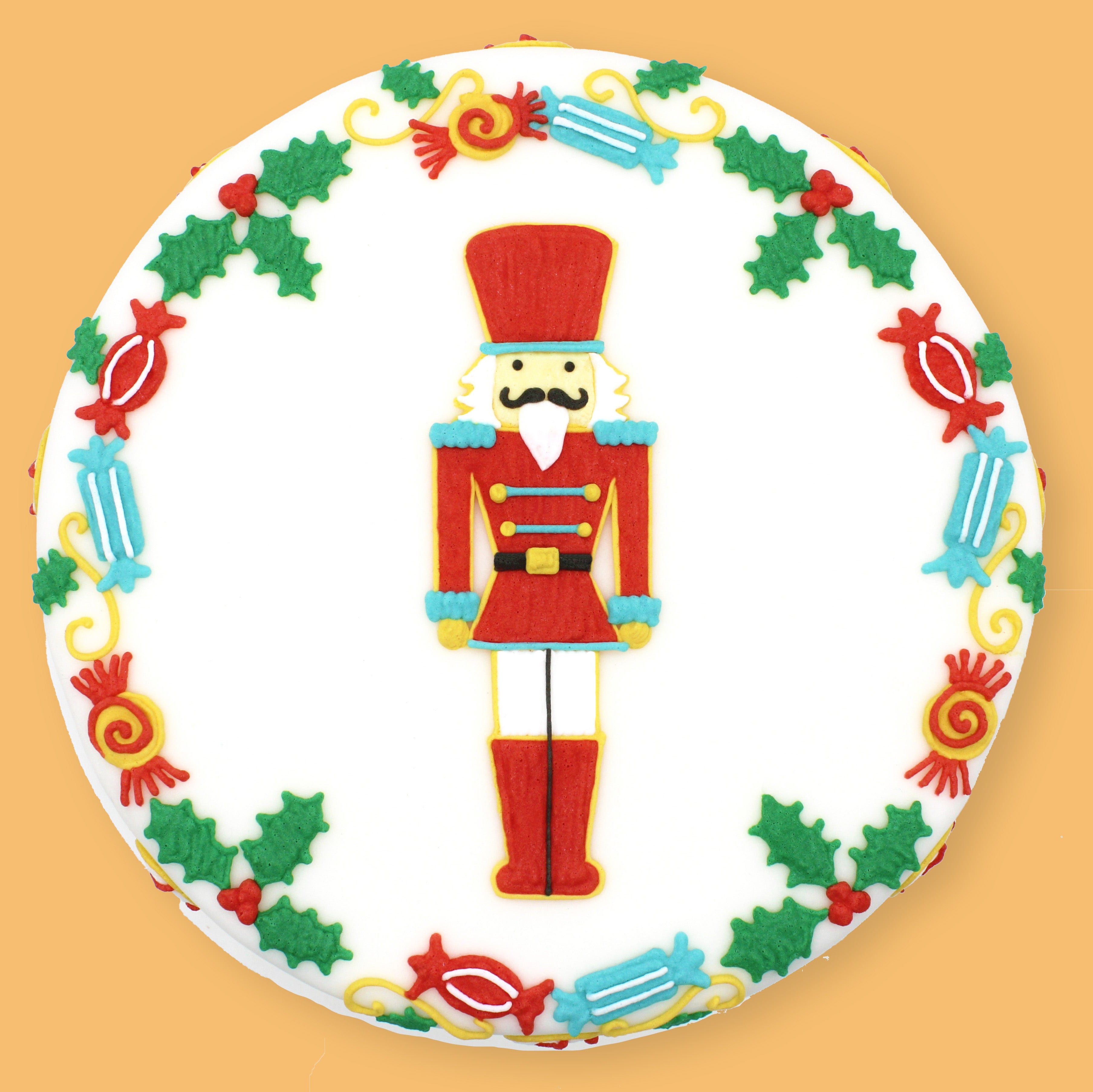 A round white cake decorated with a festive Christmas theme, featuring a central nutcracker figure and a border of holly leaves and candy motifs, set against a solid orange background.